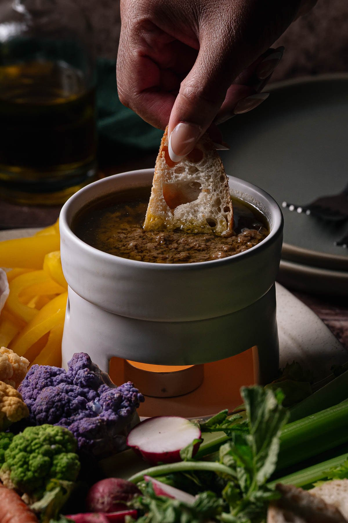 A hand dips a piece of bread into a ceramic pot of Bagna cauda, surrounded by assorted fresh vegetables and a lit candle underneath.
