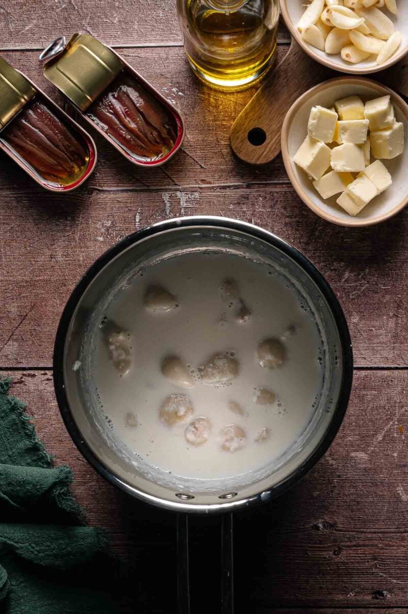 A saucepan with a milk and whole pieces of garlic; nearby are butter cubes, chopped garlic, anchovy tins, and a bottle of oil on a wooden surface.