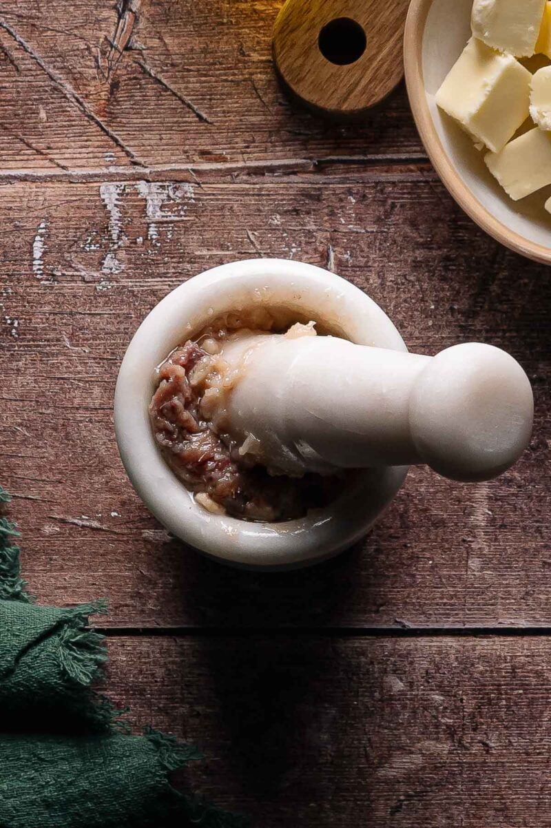 A marble mortar and pestle containing a paste, placed on a wooden surface next to a bowl of butter cubes and a green cloth.
