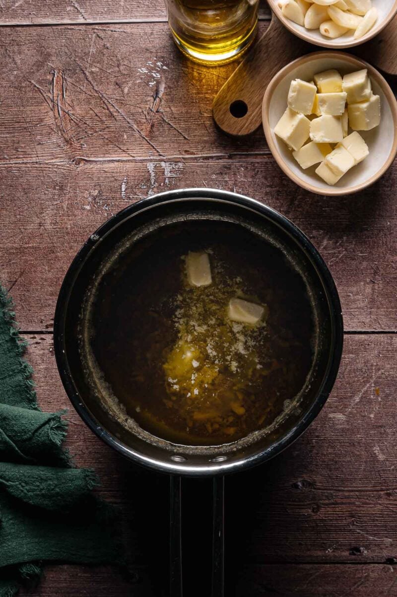 A saucepan with melting butter on a wooden surface, next to a bowl of cubed butter, garlic cloves, and a glass bottle.