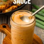 A jar of creamy bang bang sauce with a spoon, placed on a small wooden board, with green onions and food in the background.