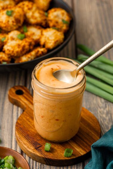 A jar of creamy bang bang dipping sauce with a spoon rests on a wooden board; a plate of breaded food and green onions are nearby.