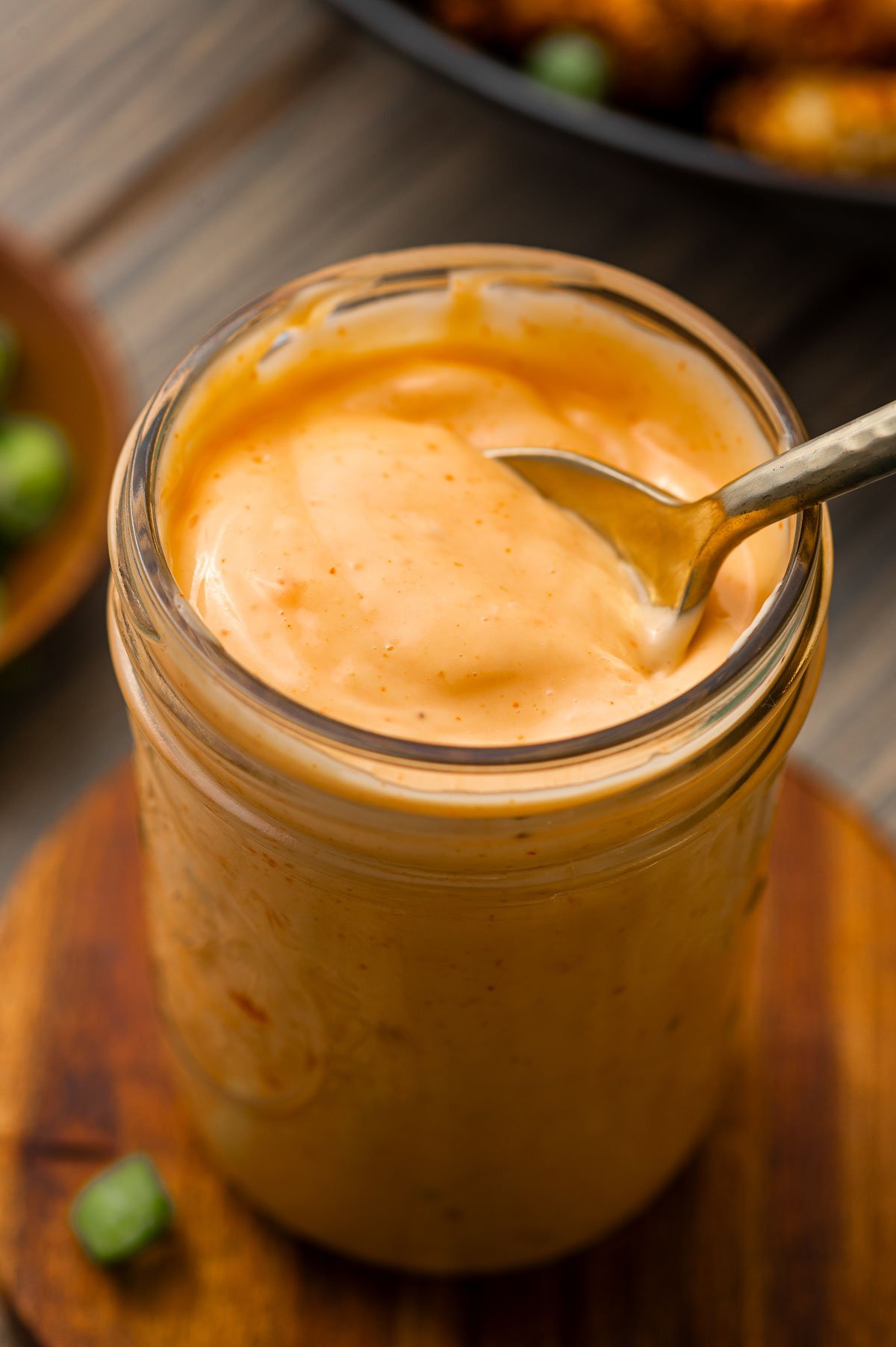 A glass jar filled with creamy, bang bang sauce. A spoon is resting inside the jar. The jar is placed on a wooden surface.