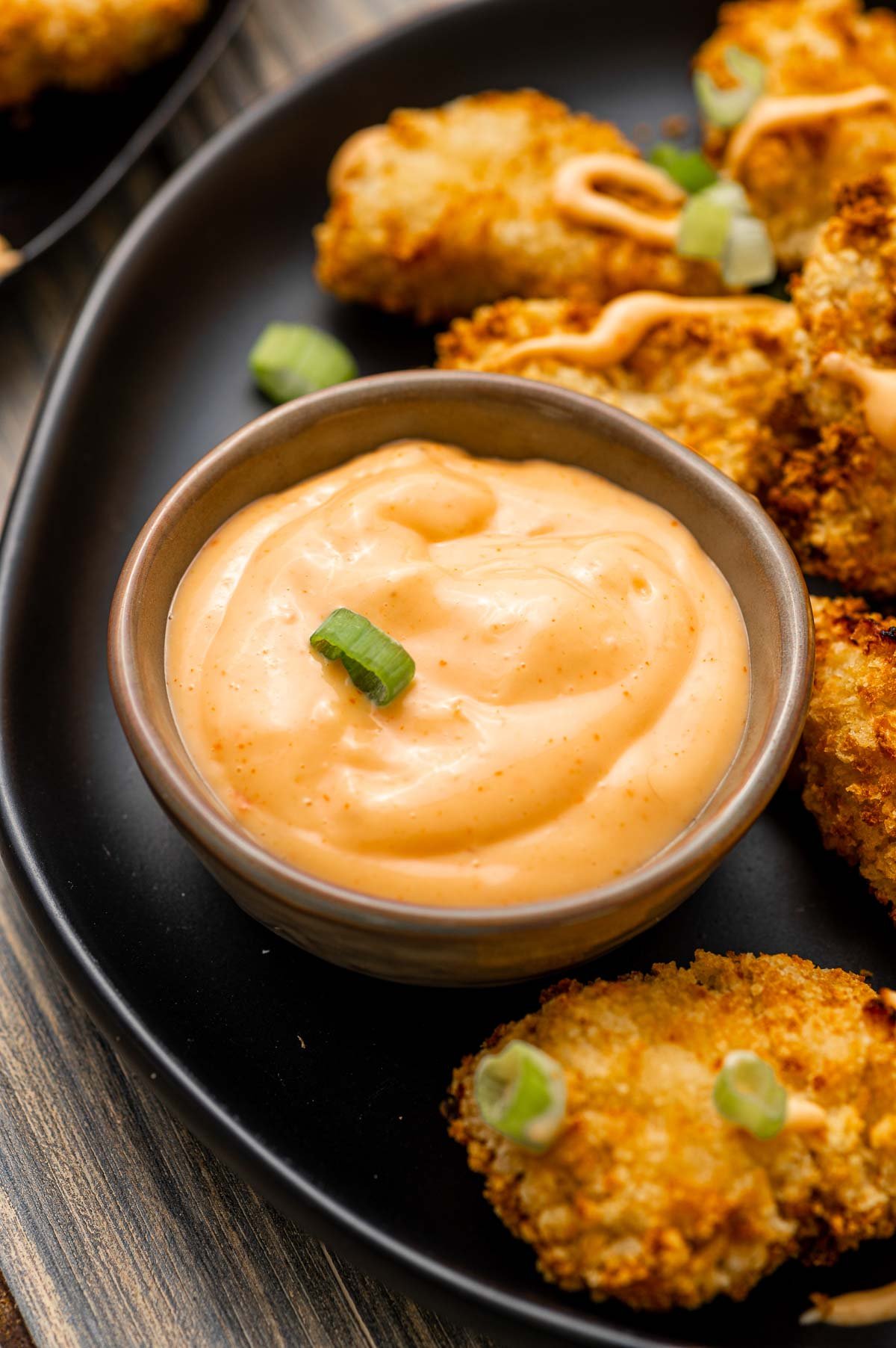 A small bowl of creamy bang bang dipping sauce garnished with chopped green onion, served on a black plate with breaded, baked appetizers.