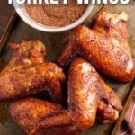 Three seasoned and smoked turkey wings on a baking sheet with a bowl of Cajun spice mix, shown from above. Text overlay reads, "How to Make Cajun Smoked Turkey Wings.