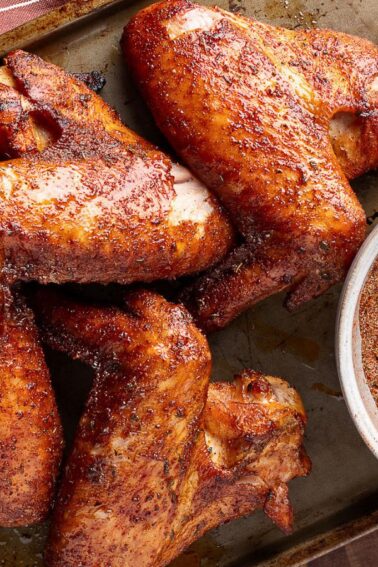 Five seasoned and cooked turkey wings on a baking tray next to a bowl of dry spice rub with a wooden spoon.