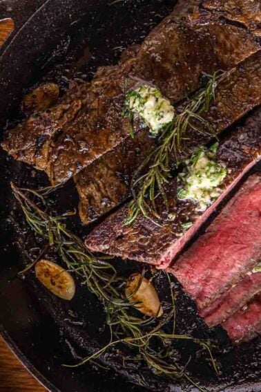 A cast iron skillet with sliced cooked steak, topped with herb butter and garnished with rosemary and garlic cloves.