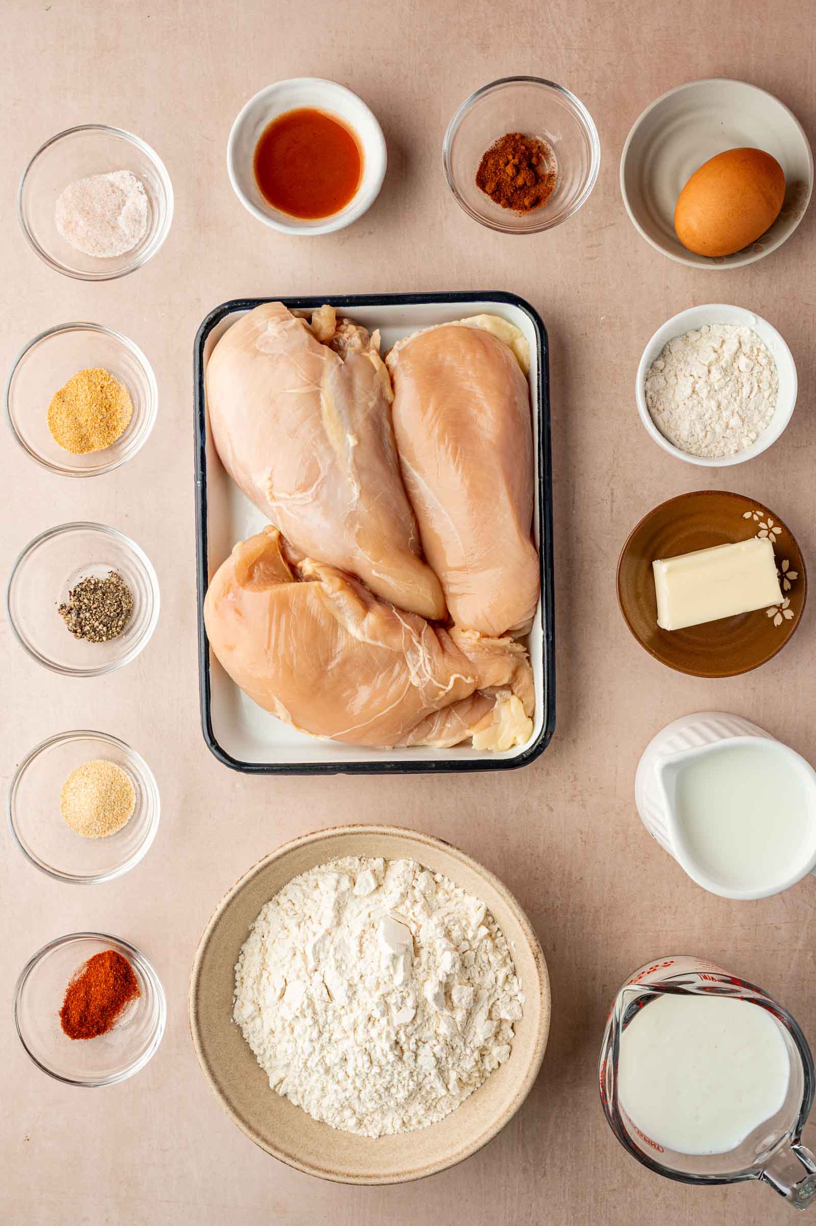 Top-down view of raw chicken breasts on a tray surrounded by small bowls containing flour, spices, butter, milk, egg, and hot sauce on a light surface.