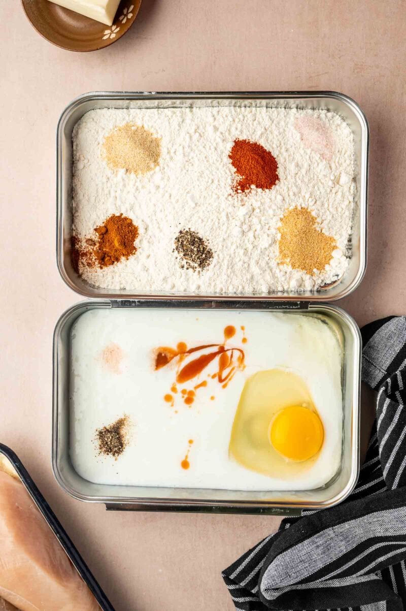 Two metal trays side by side: one contains flour and various spices, the other contains buttermilk, an egg, hot sauce, and seasonings ready for mixing.