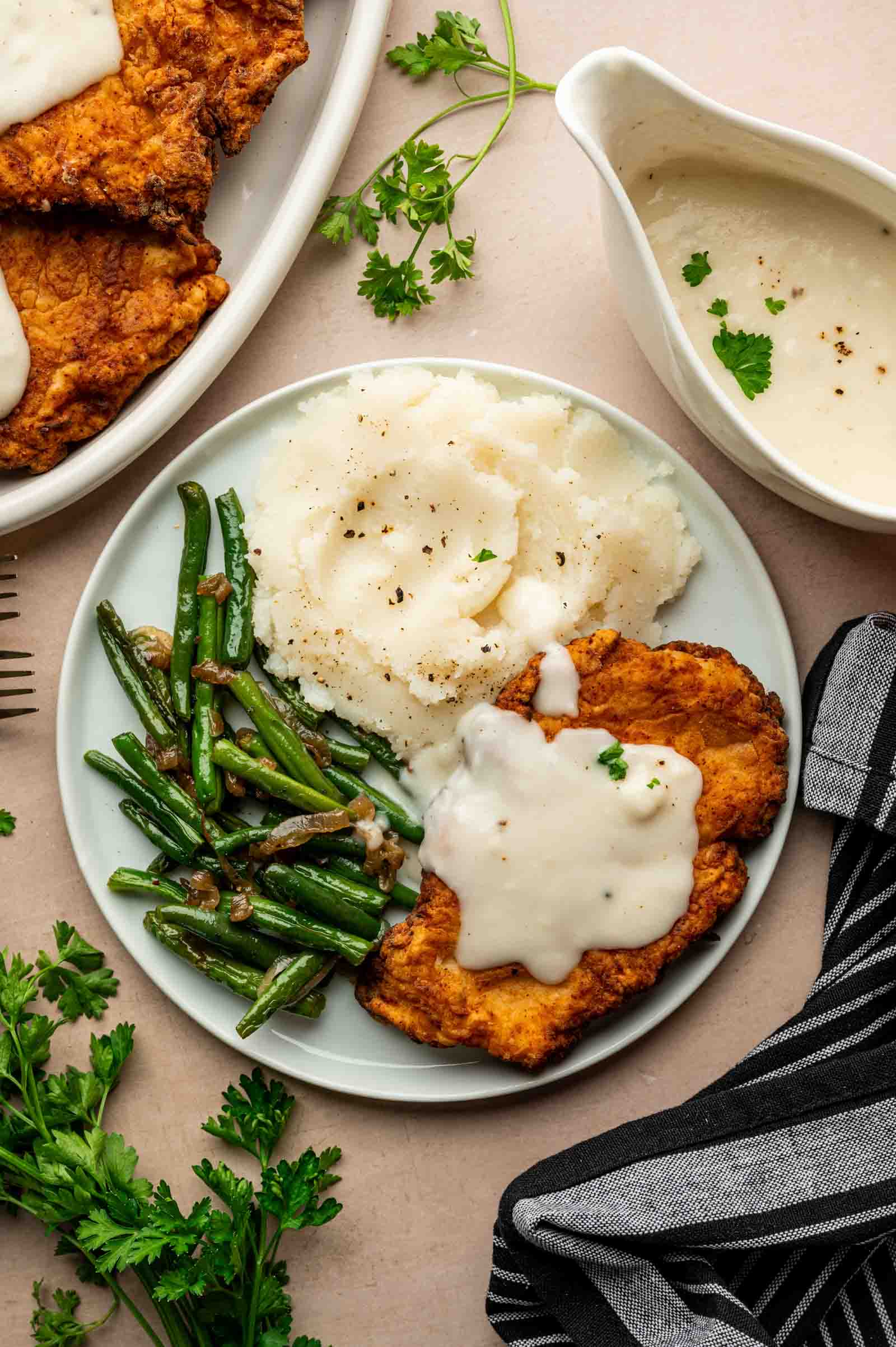 A plate with chicken fried chicken topped with white gravy, mashed potatoes, and sautéed green beans, served beside a bowl of extra gravy and fresh herbs.