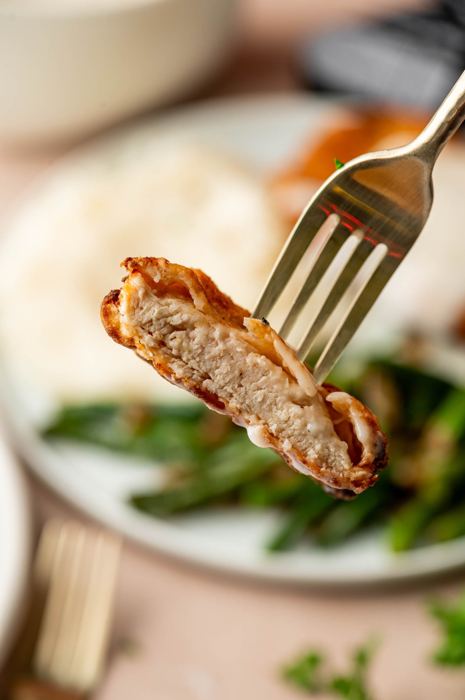 A fork holds a bite of cooked, chicken fried chicken with crispy fry coating and gravy, with green beans and potatoes blurred in the background on a plate.