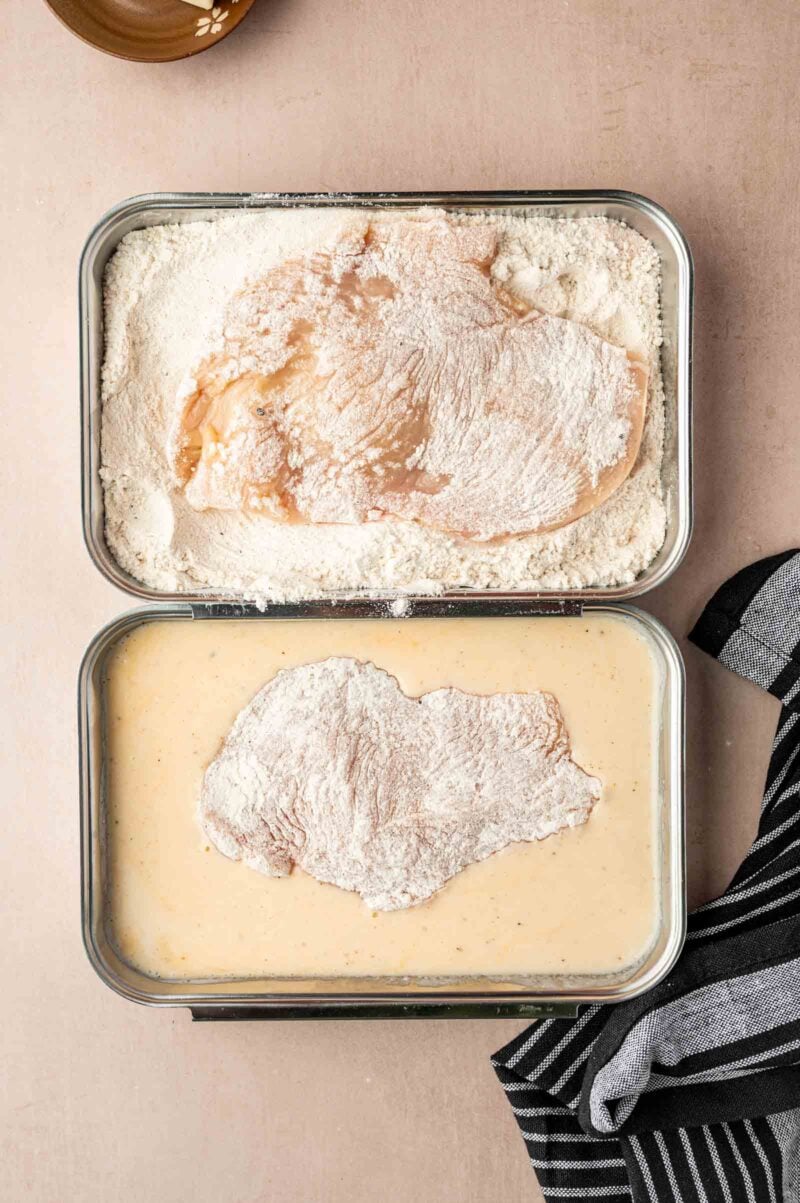 Two breading trays with raw chicken breast: one tray with chicken coated in flour, the other with chicken in a batter mixture. A striped cloth is partially visible to the right.