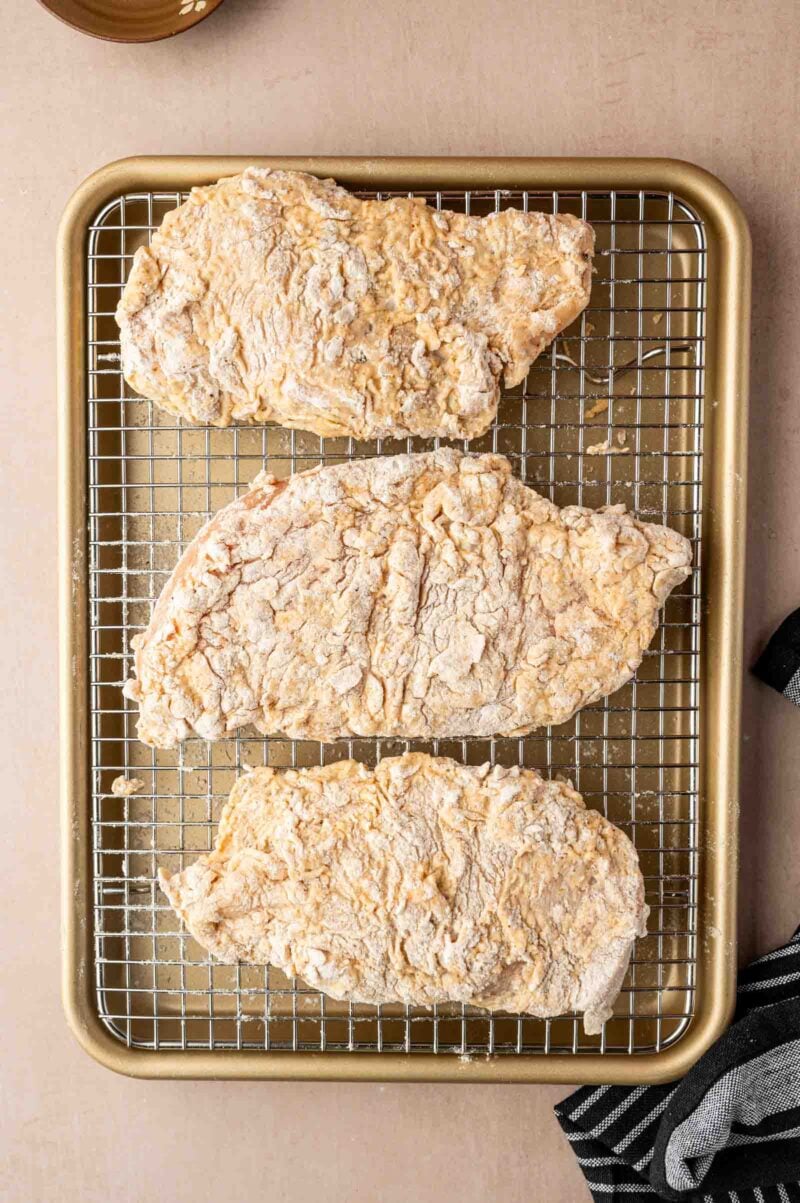 Three uncooked, breaded chicken breasts are arranged on a metal cooling rack over a gold baking sheet.