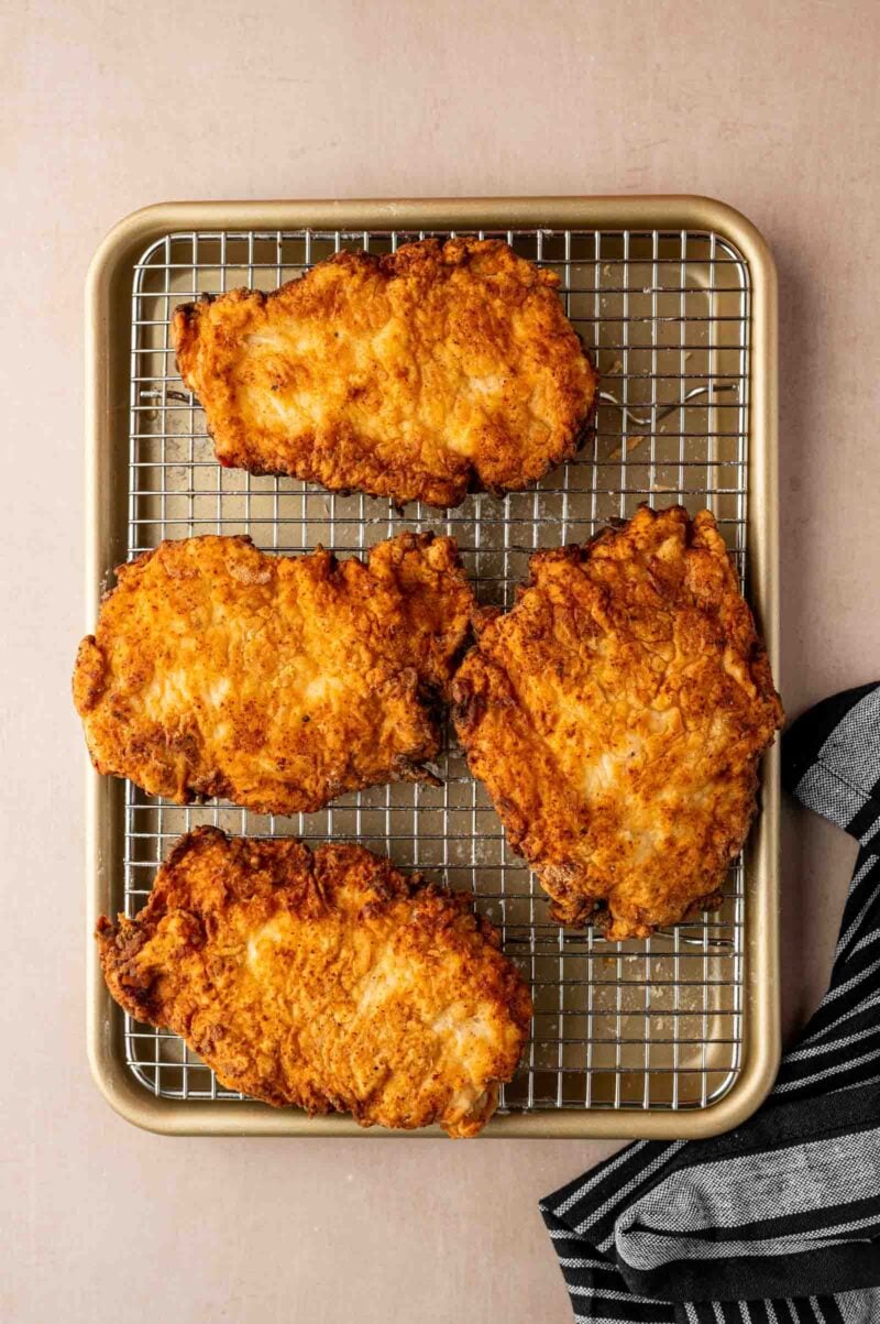Four pieces of golden-brown fried chicken on a wire rack over a baking sheet, with a striped cloth to the side.