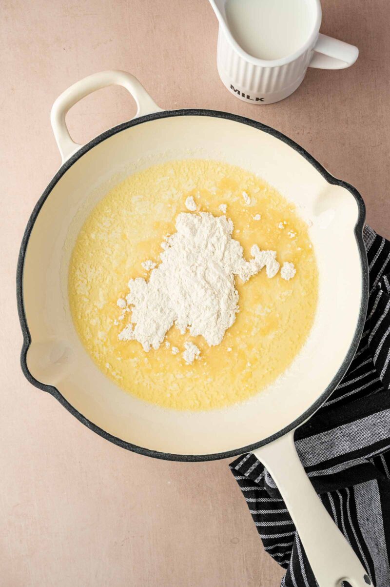 A white pan contains melted butter and a mound of flour. A striped towel is nearby, and a cup labeled "MILK" sits in the background.
