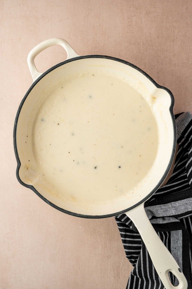 A white enamel pot filled with creamy white gravy sits on a light surface, next to a black and white striped towel.