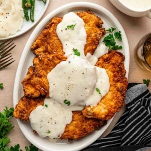 Plate of chicken fried chicken topped with white gravy, garnished with parsley, next to a striped napkin and sides of green beans and mashed potatoes.