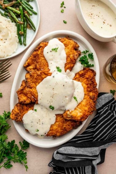 Plate of chicken fried chicken topped with white gravy, garnished with parsley, next to a striped napkin and sides of green beans and mashed potatoes.