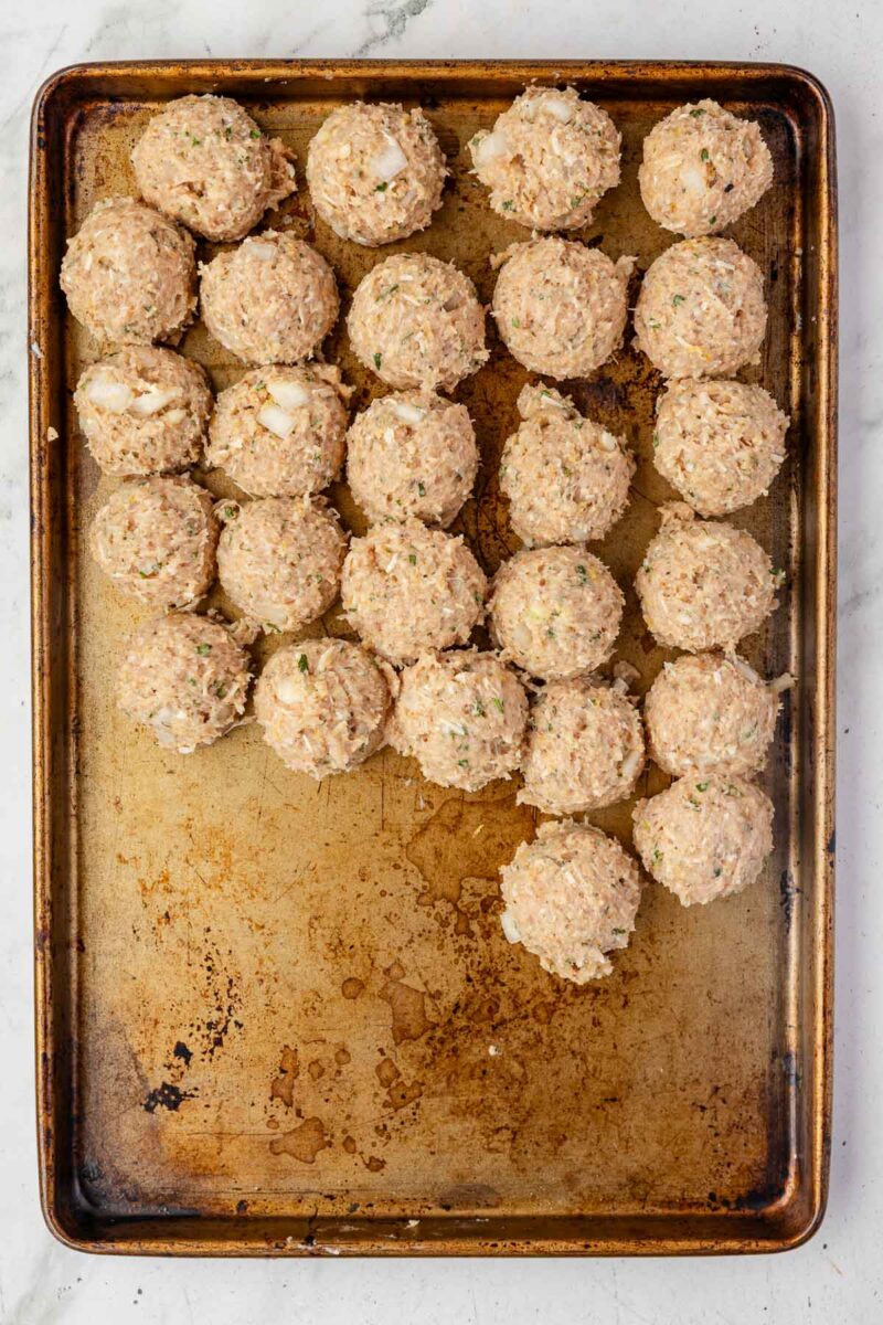 A baking sheet with evenly spaced, uncooked chicken meatballs arranged in rows on a lightly stained surface.