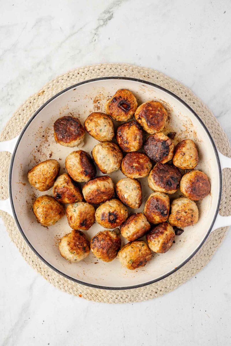 A white skillet filled with browned Chicken Piccata Meatballs sits on a round woven placemat atop a marble surface.