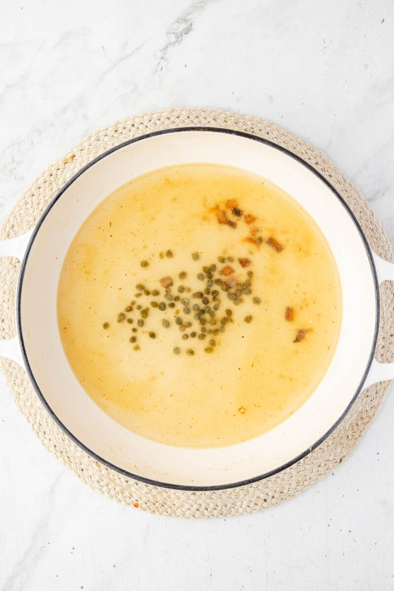 A white pot filled with a light yellow piccata sauce, garnished with green capers, sits on a woven mat atop a marble surface.