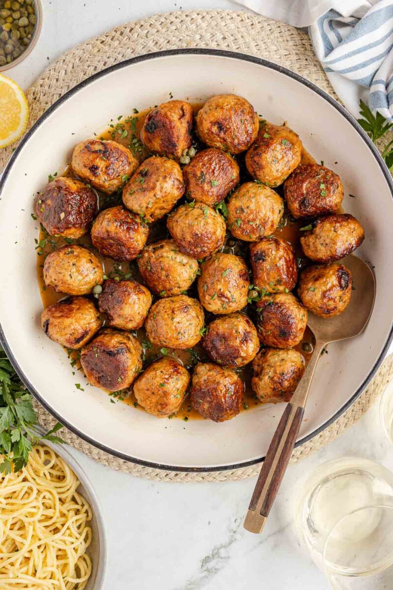 A white pan filled with browned Chicken Piccata Meatballs garnished with herbs, next to a bowl of cooked spaghetti and a glass of white wine.