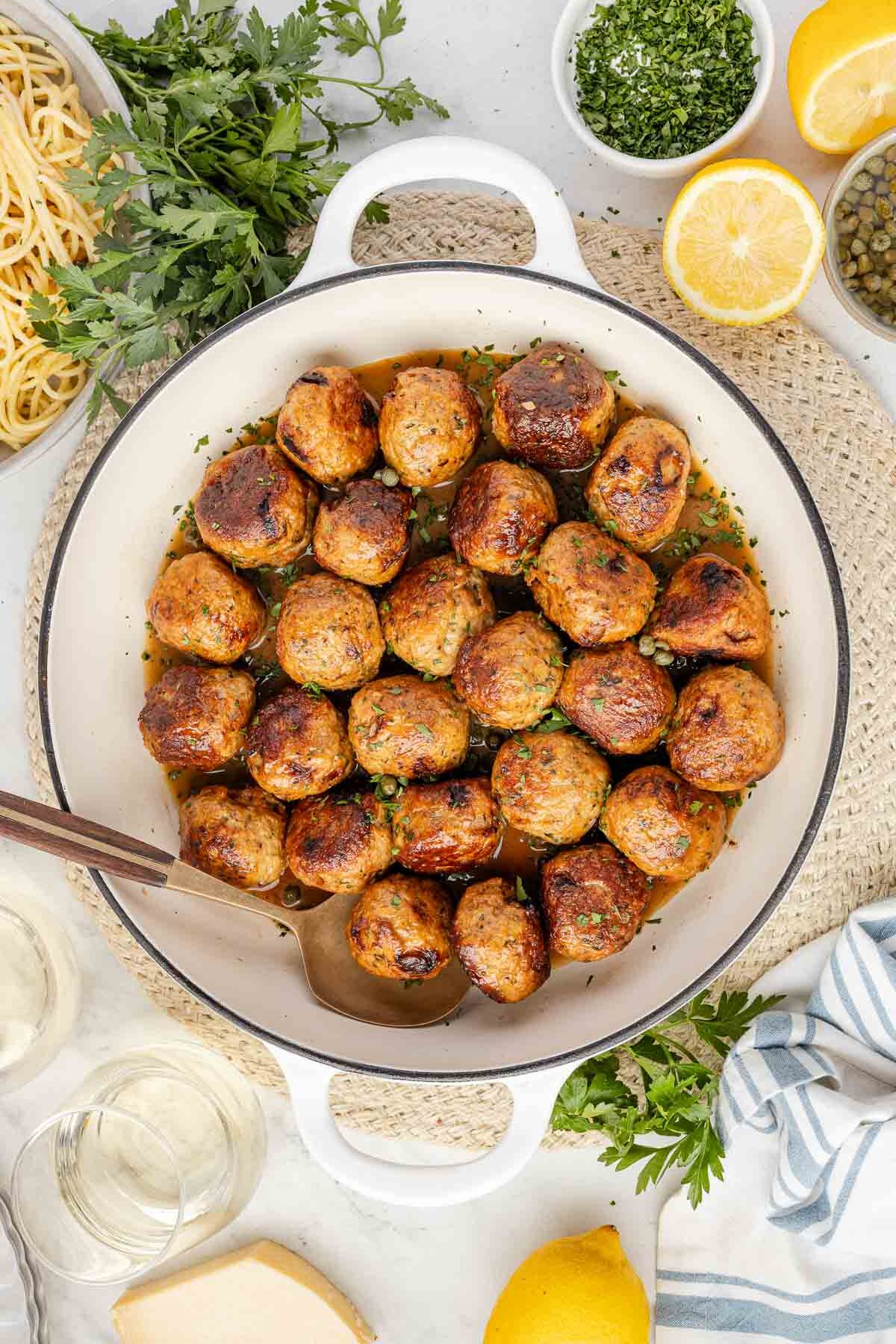 A white pot filled with browned Chicken Piccata Meatballs, garnished with herbs, on a light woven placemat, surrounded by spaghetti, lemon halves, parsley, capers, and a glass of white wine.