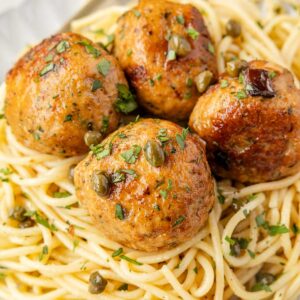 Four browned Chicken Piccata Meatballs garnished with herbs and capers served on a bed of spaghetti on a white plate.