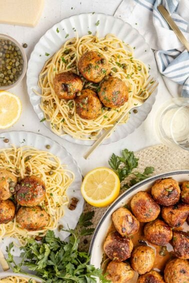 Plates of spaghetti topped with browned Chicken Piccata Meatballs, garnished with herbs and lemon slices, with a skillet of meatballs and fresh parsley nearby.