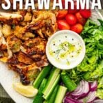Plated chicken shawarma with sliced cucumbers, lettuce, cherry tomatoes, red onions, lemon wedges, and a side of white sauce. Text overlay: "How to Make Chicken Shawarma.