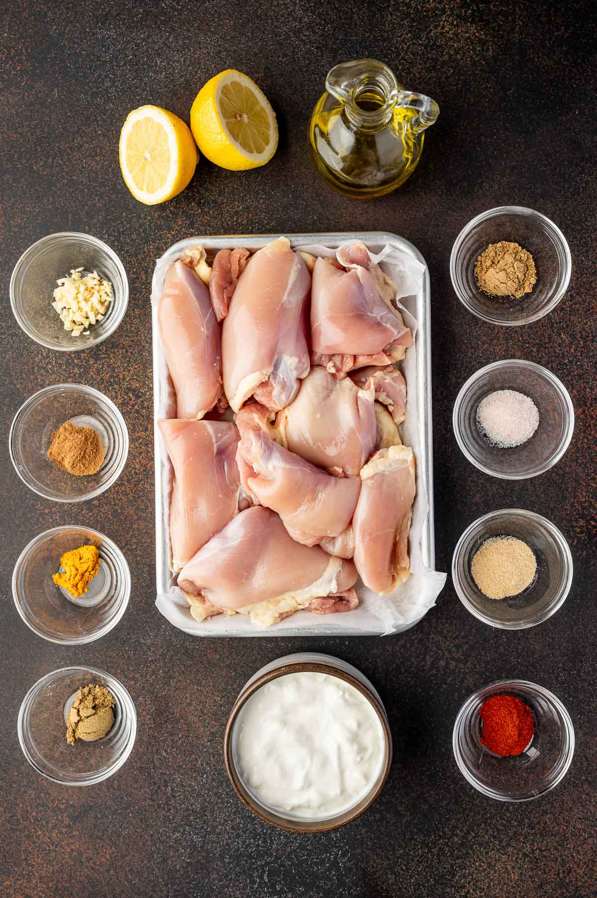 Raw chicken thighs in a tray surrounded by small bowls of spices, minced garlic, yogurt, lemon halves, and a bottle of olive oil on a dark surface.