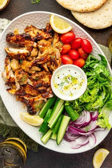 A platter with chicken shawarma, sliced cucumbers, cherry tomatoes, lettuce, red onion, lemon wedges, flatbread, and a bowl of yogurt sauce.