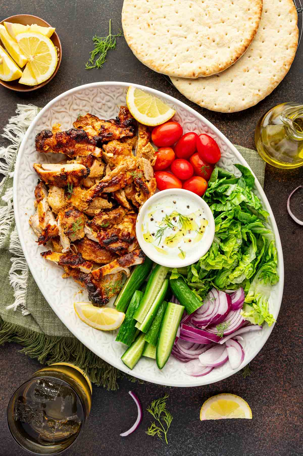 Oval platter with chicken shawarma, lettuce, cucumber, cherry tomatoes, red onion, lemon wedges, flatbread, and a bowl of white dipping sauce on a dark surface.