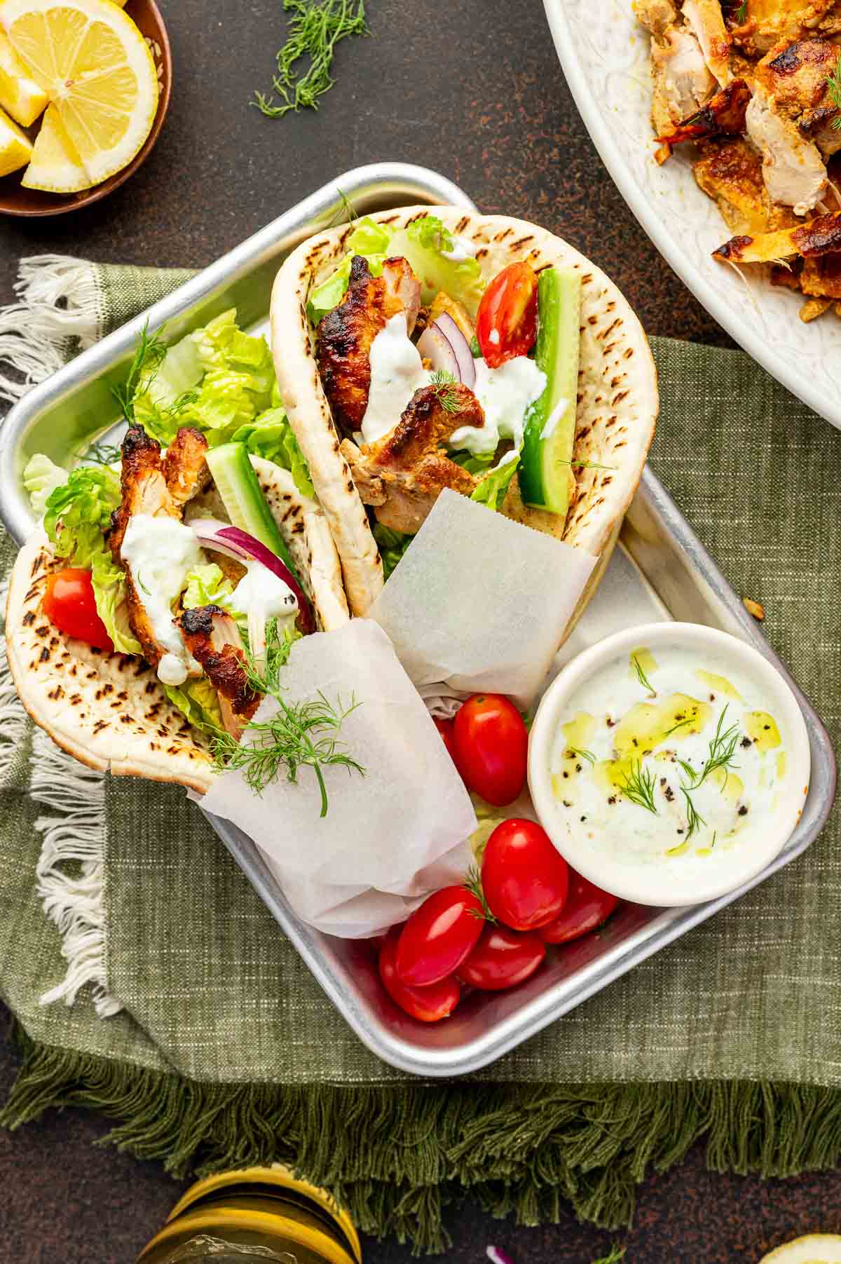 Two chicken shawarma with lettuce, tomato, cucumber, onion, and tzatziki in pita bread, served on a tray with cherry tomatoes and a small bowl of tzatziki sauce.