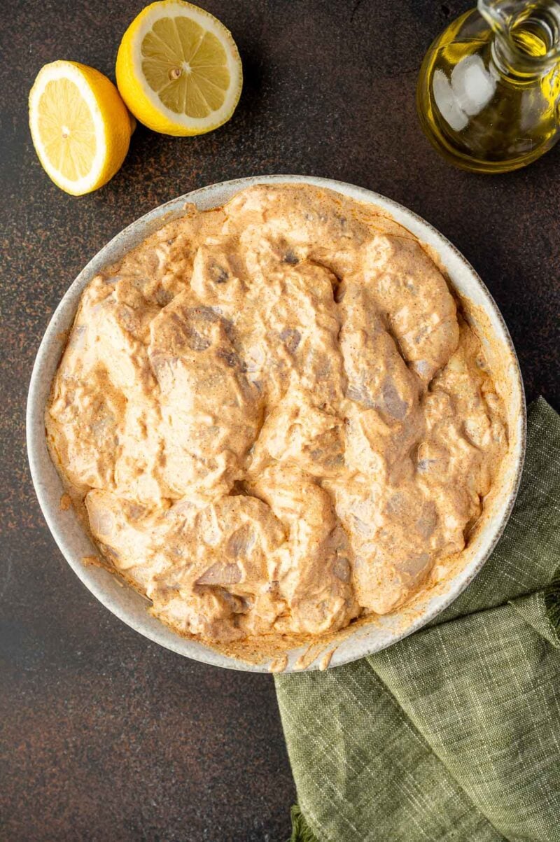 A bowl of marinated chicken pieces in a creamy, spiced sauce, next to a halved lemon, a bottle of oil, and a green cloth on a dark surface.