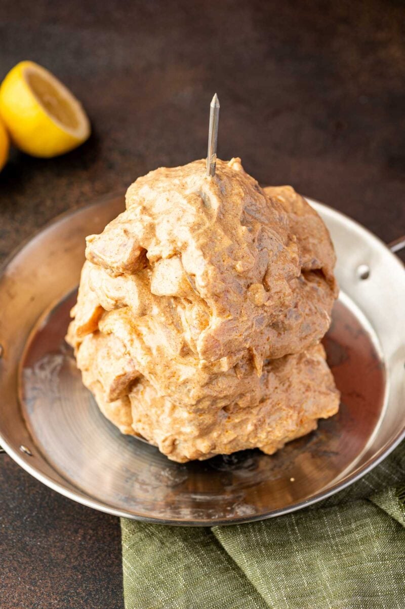 A stack of marinated raw chicken pieces is skewered vertically on a metal rod in a round metal pan, with a halved lemon in the background.