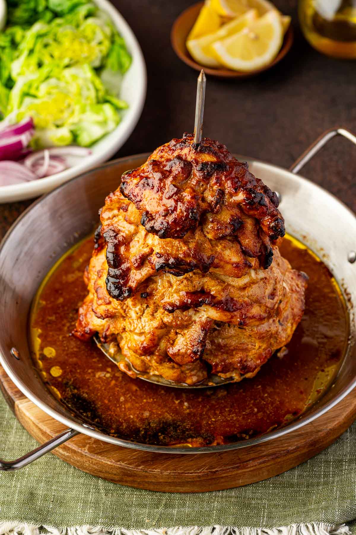 A stack of marinated chicken shawarma slices roasted on a vertical spit is served in a metal pan with juices, with salad and lemon wedges in the background.