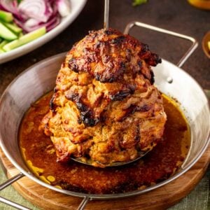 A stack of marinated and roasted chicken shawarma on a vertical spit in a round metal pan, with salad ingredients visible in the background.