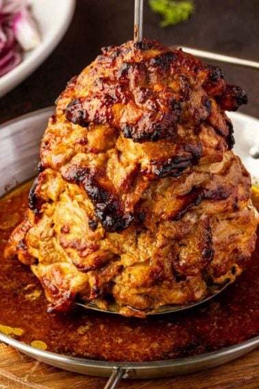 A stack of seasoned, grilled chicken shawarma sits upright in a round metal pan with juices, surrounded by lemon wedges, sliced vegetables, and pita on the table.