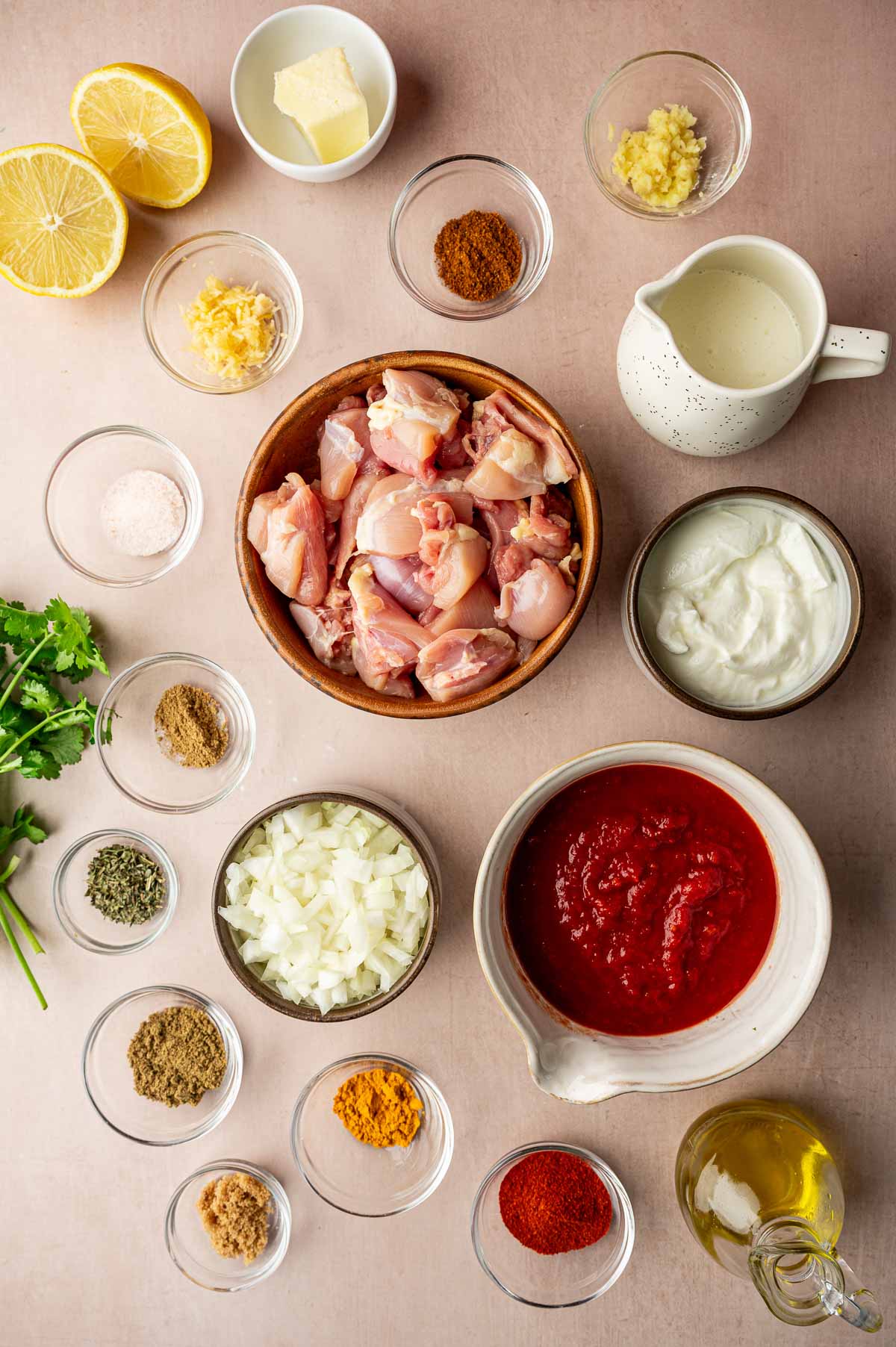 A flat lay of butter chicken tikka masala ingredients including raw chicken pieces, tomato sauce, yogurt, cream, spices, chopped onions, garlic, ginger, lemon, butter, and fresh cilantro arranged in bowls.