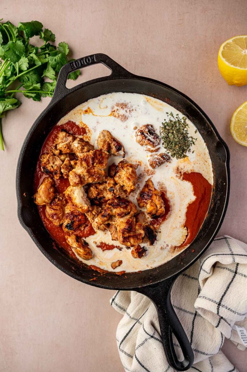 A cast iron skillet contains cooked chicken pieces, tomato sauce, cream, and dried herbs. Fresh cilantro and halved lemons are on the side, with a dish towel nearby.