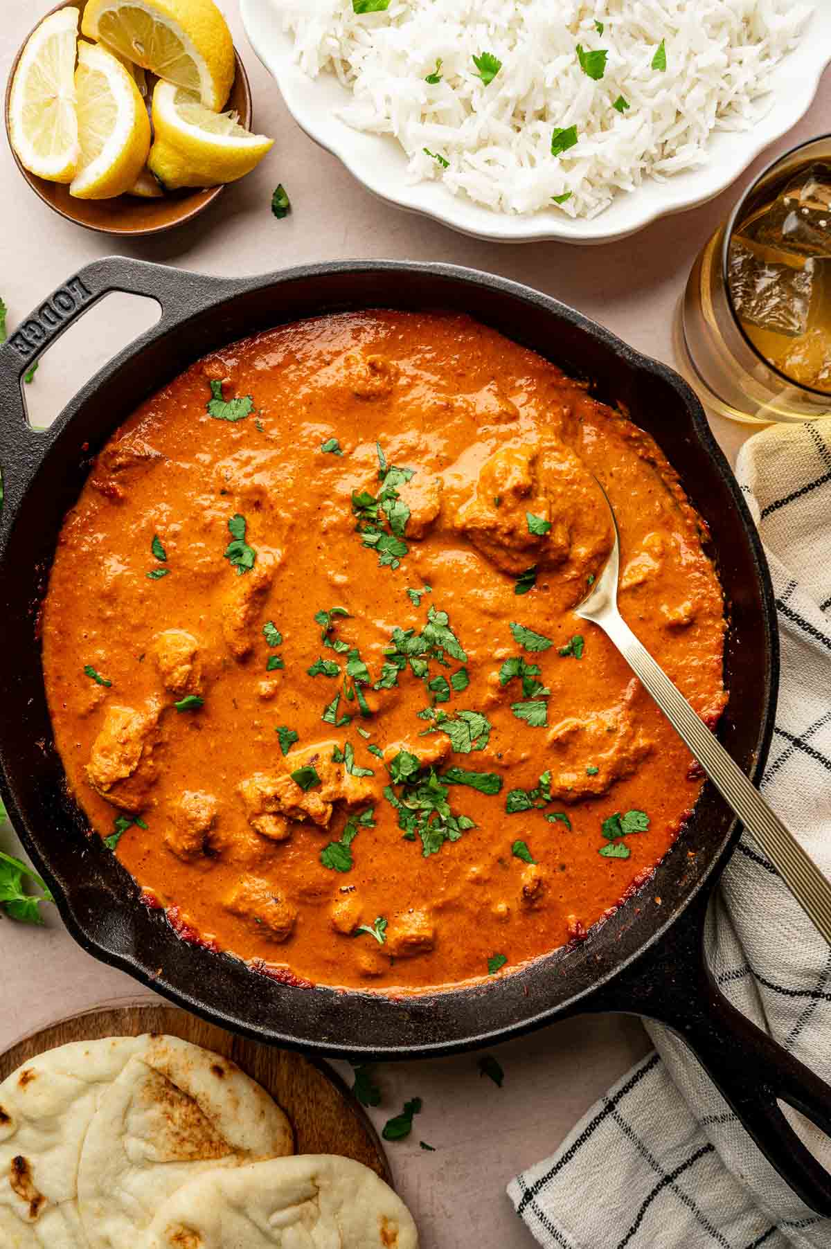 A cast iron skillet filled with chicken tikka masala garnished with cilantro, next to naan bread, a bowl of white rice, lemon wedges, and a glass of iced drink.