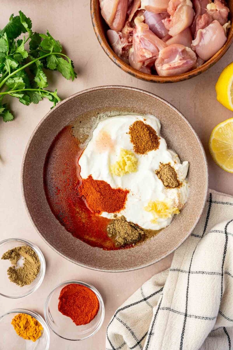 A bowl with yogurt, ground spices, and minced garlic, next to raw chicken pieces, fresh cilantro, lemon halves, and a kitchen towel.