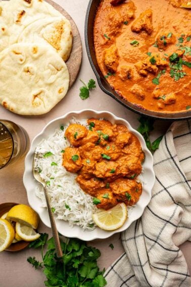 Plate of chicken tikka masala with white basmati rice garnished with cilantro and lemon, naan bread on the side, and a skillet of curry, with a drink and napkin nearby.