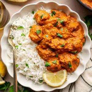 A plate with white rice and chicken tikka masala garnished with chopped herbs and a lemon slice, served with naan bread on the side.