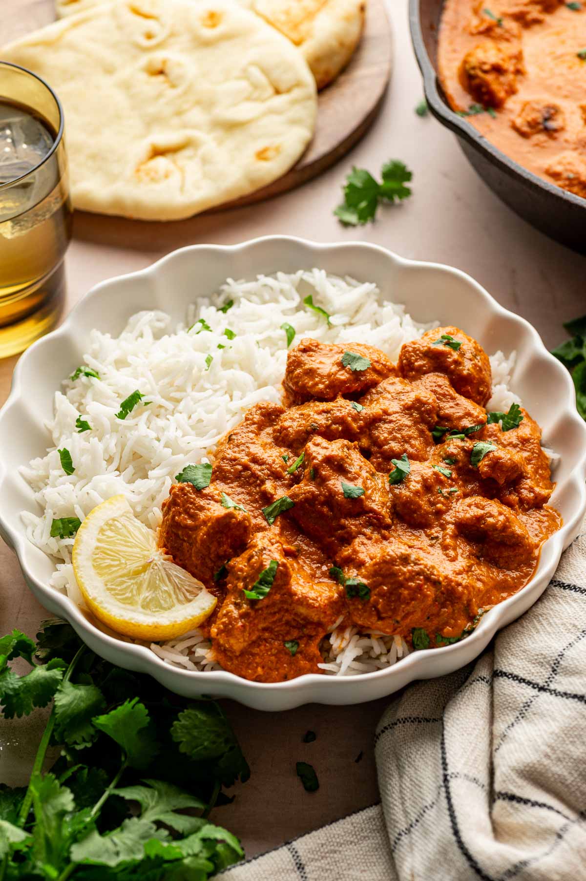 A bowl of white rice topped with chicken tikka masala, garnished with cilantro and a lemon wedge, with naan bread and a glass of drink in the background.