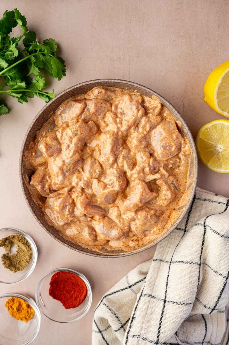 A bowl of marinated chicken pieces in a creamy orange sauce, surrounded by fresh cilantro, lemon halves, a cloth napkin, and small bowls of ground spices.