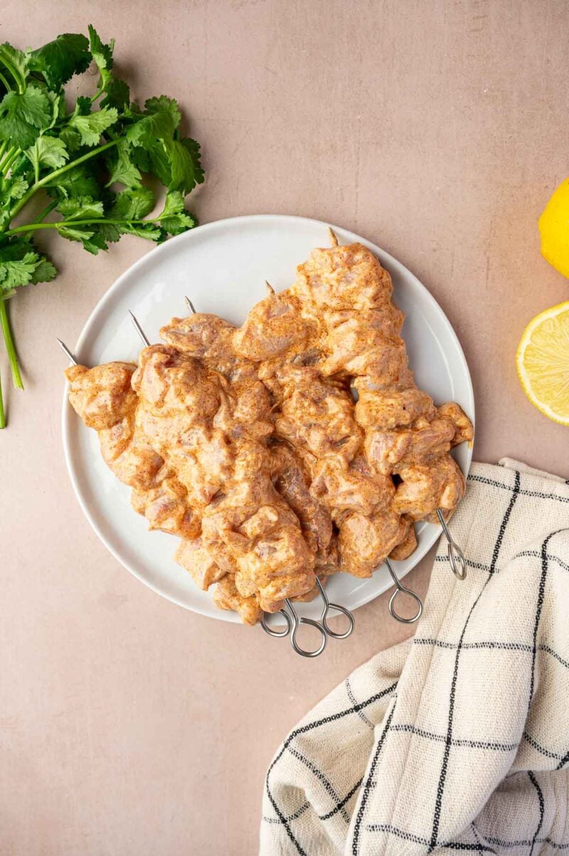 Plate of marinated chicken skewers on a white dish, with a checkered cloth, cilantro, and half a lemon on a beige surface.