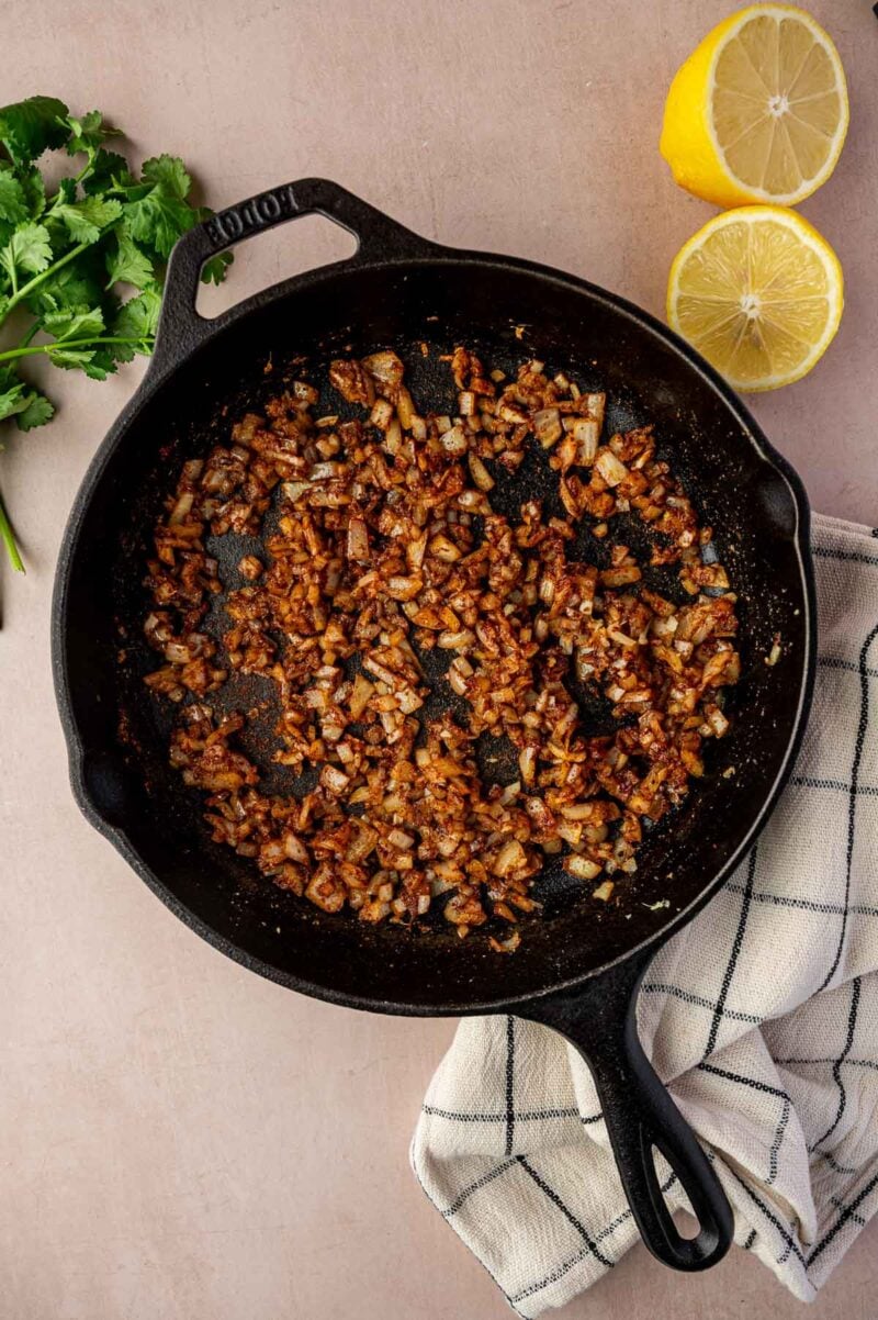 A cast iron skillet with sautéed onions and spices sits on a cloth, next to a bunch of cilantro and a halved lemon on a light surface.