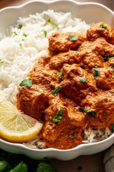 A plate of basmati rice topped with chicken tikka masala and garnished with chopped cilantro, served with lemon wedges on the side.
